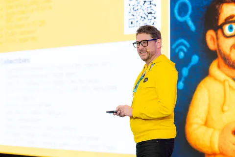 A speaker mid-talk on the Quality Land 2026 stage, an illustrated portrait and a QR code on the slide behind him.